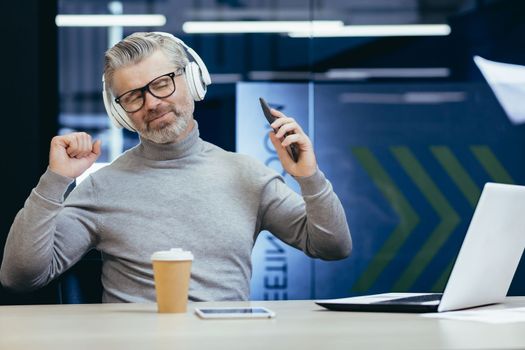 Portrait of senior gray-haired man listening to music in headphones, sitting at desk in office, holding phone with eyes closed, dancing, relaxed.