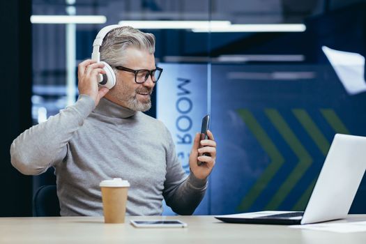 Senior gray haired businessman boss working inside modern office using laptop at work, mature man in headphones listening to music and audiobooks podcasts using app on phone.