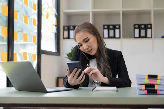 Smiling beautiful Asian businesswoman analyzing chart and graph showing changes on the market and holding smartphone at office...