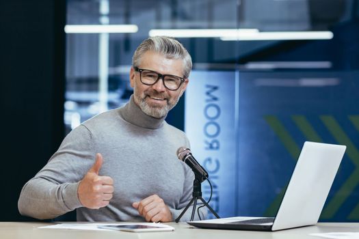 Portrait of a handsome senior man sitting in an office, studio at a table with a microphone. Record podcast, interview, record audio. A smiling man looks at the camera. He points a super finger.