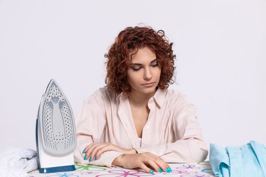 The girl sits at the ironing board, quite tired of this work. Daily housework after washing clothes is ironing shirts.
