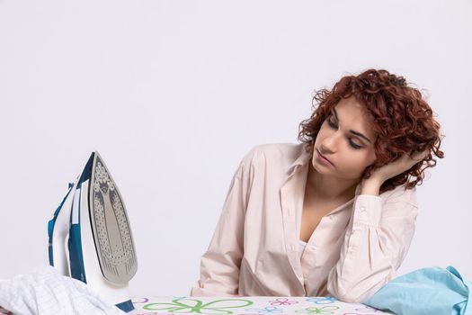 The girl sits at the ironing board, quite tired of this work. Daily housework after washing clothes is ironing shirts.