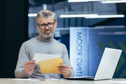 Upset senior man businessman sitting in the office at the desk, holding an envelope with a letter in his hands, received bad news, bank account for a loan, debt.