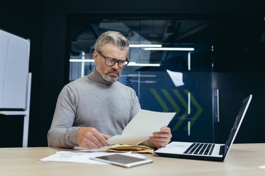 Mature gray-haired businessman inside modern office received letter message, senior man upset opens postal envelope, boss reads bad news upset and sad working at desk with laptop.