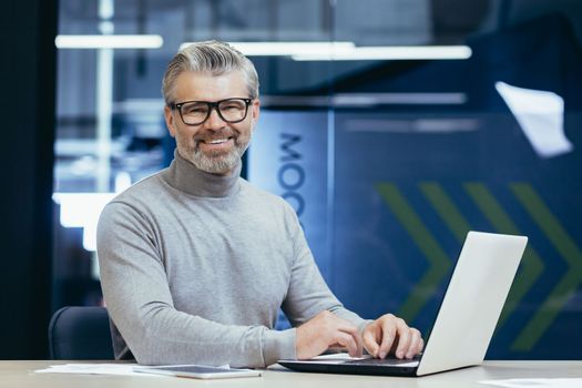Portrait of senior man inside office, mature businessman with beard smiling and looking at camera, boss working at desk using laptop at work.