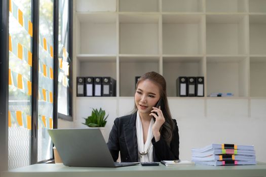 Smiling beautiful Asian businesswoman analyzing chart and graph showing changes on the market and holding smartphone at office...