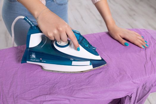 A woman irons a purple men's shirt with an iron on an ironing board. Close-up view. A woman's hand with nails in blue with glitter.