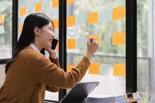 Smiling young asian woman writing on sticky notes on window in creative office.