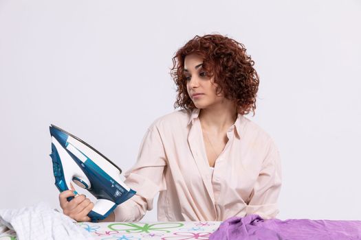 A young girl sits at the ironing board and iron. Curly short and dark hair. A stand for ironing clothes.
