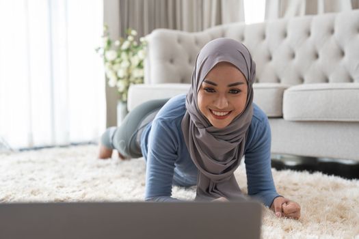 Asian young woman is practicing yoga in the livingroom. A Muslim woman is looking at a video lesson of yoga on her laptop and exercising at home. Fit arabic woman training at home.