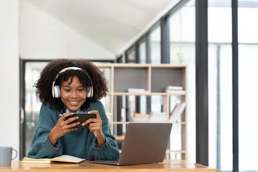 Photo of cheerful joyful mixed race woman playing games and wearing wireless headphones at home Play game on your smartphone that wins the moment of victory. Fun and enjoyable vacation.