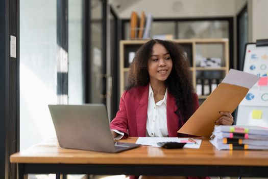 Smiling young African american female Finance Entrepreneur sitting in workplace office hoding coffee cup and working on a laptop, investment financial document report finance analysis concept
