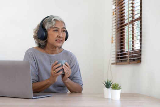 Senior woman using laptop while wearing headphones at home - Joyful elderly lifestyle and technology concept.