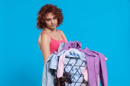 Woman carries a basket full of ironing shirts. A figure isolated from a solid blue background. A bored expression on her face. Dark curly hair.