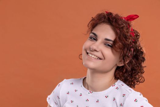 European beauty girl with short and curly hair. Uniform background in brick-red color. She is wearing a white blouse with a cherry pattern.