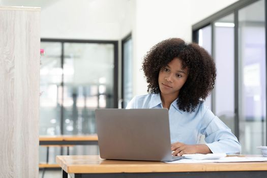 Smiling young African female entrepreneur sitting at a desk in her home office working online with a laptop.