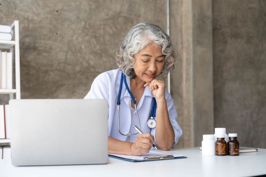 Portrait of senior grey-haired female doctor in her office using laptop for video chat with a patient. Online consultation with doctor for diagnoses and treatment recommendation