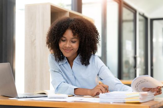 Smiling young African female entrepreneur sitting at a desk in her home office working online with a laptop.
