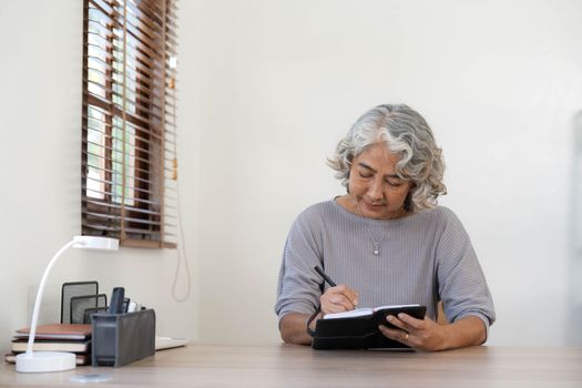 Senior stylish woman taking notes in notebook while using laptop at home