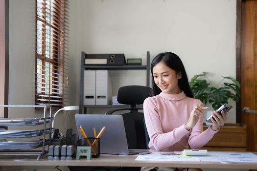 Smiling beautiful Asian businesswoman analyzing chart and graph showing changes on the market and holding smartphone at office...
