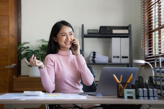 Smiling beautiful Asian businesswoman analyzing chart and graph showing changes on the market and holding smartphone at office...