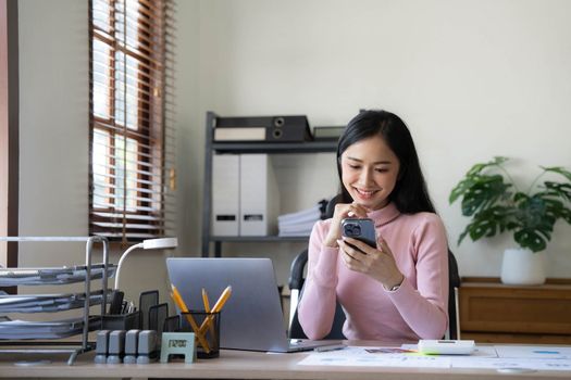 Smiling beautiful Asian businesswoman analyzing chart and graph showing changes on the market and holding smartphone at office...
