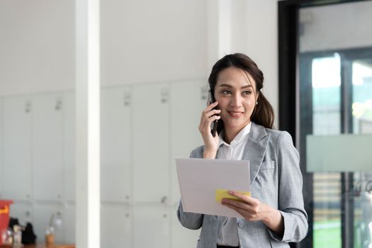 Happy young asian business woman wearing suit holding mobile phone standing in her workstation office. 