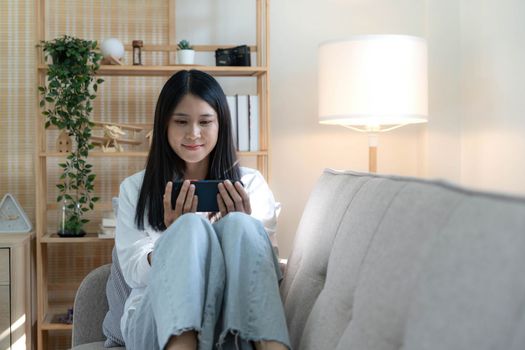 Beautiful young asian women play mobile game sitting on sofa in living room. Playing game on smartphone winning victory moment. Very enjoy and fun relax time..