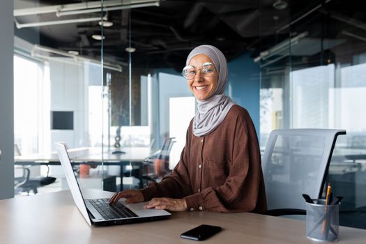 Portrait of successful businesswoman inside office with laptop, woman in hijab smiling and looking at camera, muslim office worker wearing glasses.
