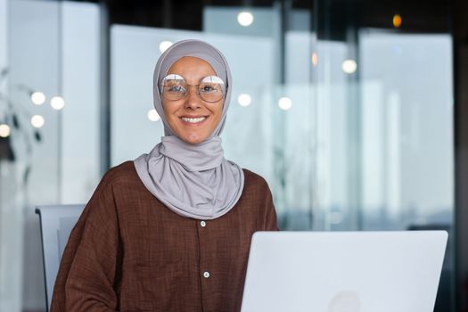 Portrait of successful businesswoman inside office with laptop, woman in hijab smiling and looking at camera, muslim office worker wearing glasses.