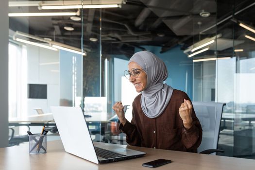 Successful businesswoman boss celebrating victory and successful triumph, muslim in hijab looking at laptop screen and holding hand up gesture of success and achievement, woman working inside