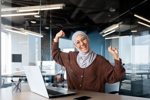 Portrait of successful businesswoman in hijab, muslim woman working inside office using laptop at work looking at camera and smiling happy, female worker celebrating victory good achievement results.