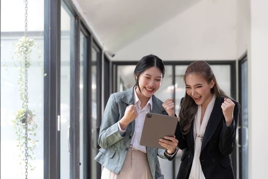 Two beautiful Asian businesswomen standing using digital tablet consulting and analyzing information in office work...