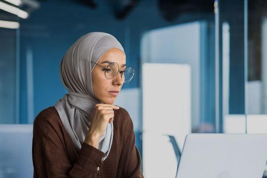 Businesswoman thinking and sad, looking at laptop screen close up, female worker in glasses and hijab working inside modern office building.