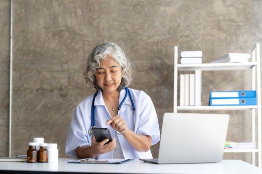 doctor senior grey-haired woman in white medical coat is using a smartphone and smiling while sit use laptop at office.