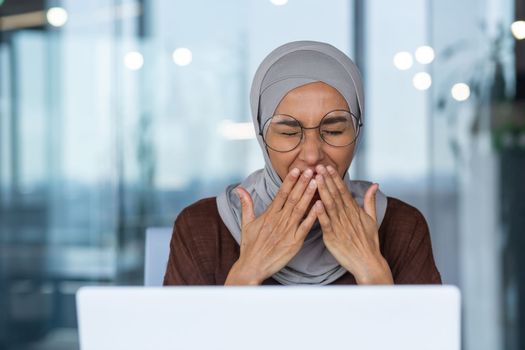 Close-up photo of businesswoman in hijab tired working with laptop yawning sitting on chair inside modern office building.