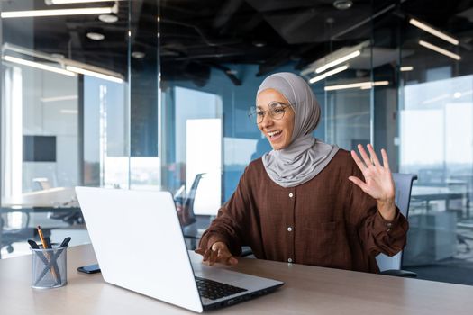 Businesswoman in hijab works inside a modern office building, Muslim talks on video call, online meeting with colleagues and clients, woman cheerfully waves her hand and greets interlocutors online.