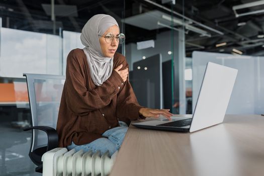 Businesswoman freezing in office at work, woman in hijab near heater trying to keep warm working inside office building with laptop, cold in office.