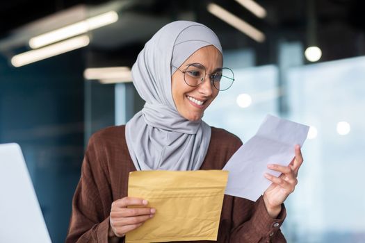 Businesswoman in hijab close up reading notification, Muslim woman received postal envelope letter with message joyfully reading and smiling, successful woman at work inside office.