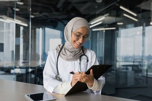A young beautiful Arab female doctor, a Muslim woman, sits in a hijab with a stethoscope in the office at the table. Works with documents, records patients' medical history.