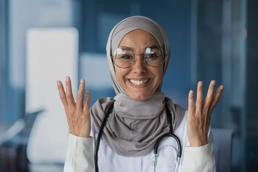 Happy and friendly young beautiful woman doctor student Muslim, Arabic. Standing in hijab, glasses and with stethoscope, looking at camera, pointing with hands.