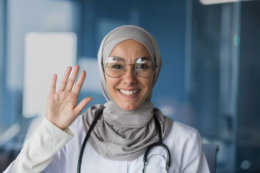 Portrait of a young beautiful woman, a Muslim, Arab woman doctor. She stands dressed in a hijab, glasses and with a stethoscope, looks and waves at the camera, smiles, greets.