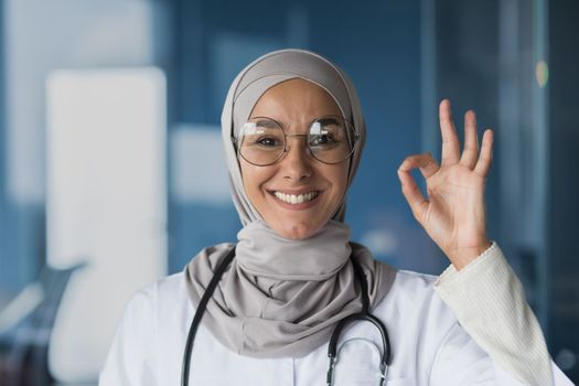 Portrait of a young, beautiful woman, an Arab, Muslim doctor. She stands dressed in a hijab, glasses and with a stethoscope, looks at the camera, smiles, points with her hand ok.