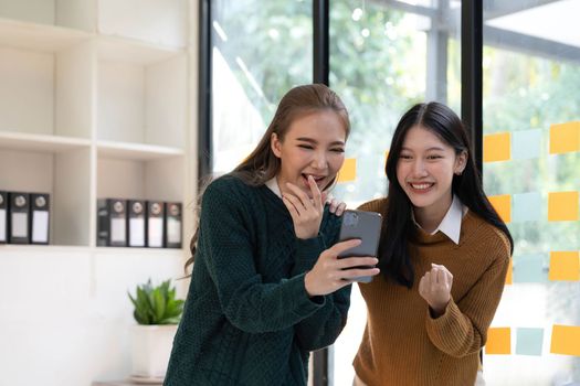 Two young Asian businesswomen show joyful expression of success at work smiling happily with a laptop computer and smartphone in a modern office...