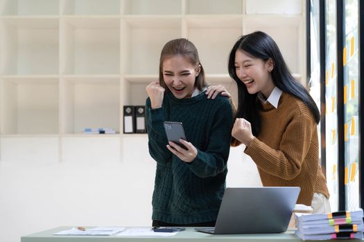 Two young Asian businesswomen show joyful expression of success at work smiling happily with a laptop computer and smartphone in a modern office...