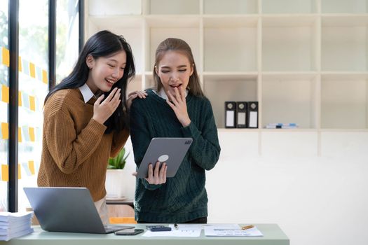 Two young Asian businesswomen show joyful expression of success at work smiling happily with a laptop computer and smartphone in a modern office...