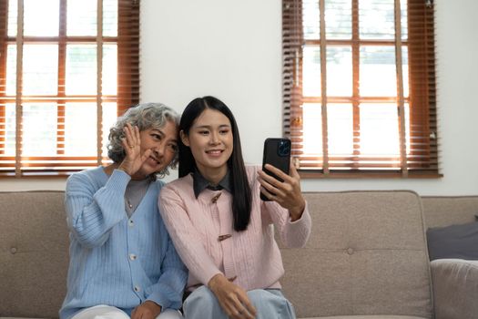 Smiling older mother and adult daughter using phone together, sitting on cozy sofa at home, happy young woman and mature mum looking at smartphone screen, watching video, having fun, two generations..