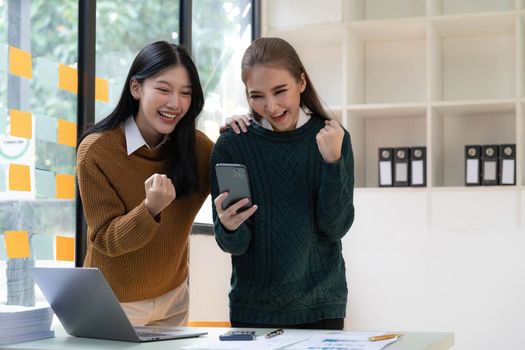 Two young Asian businesswomen show joyful expression of success at work smiling happily with a laptop computer and smartphone in a modern office...