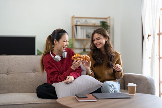 Two friends enjoying eating croissant sitting on a couch in the living room at home.