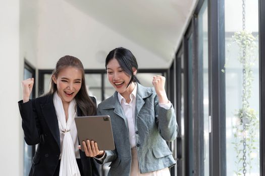 Two beautiful Asian businesswomen standing using digital tablet consulting and analyzing information in office work...